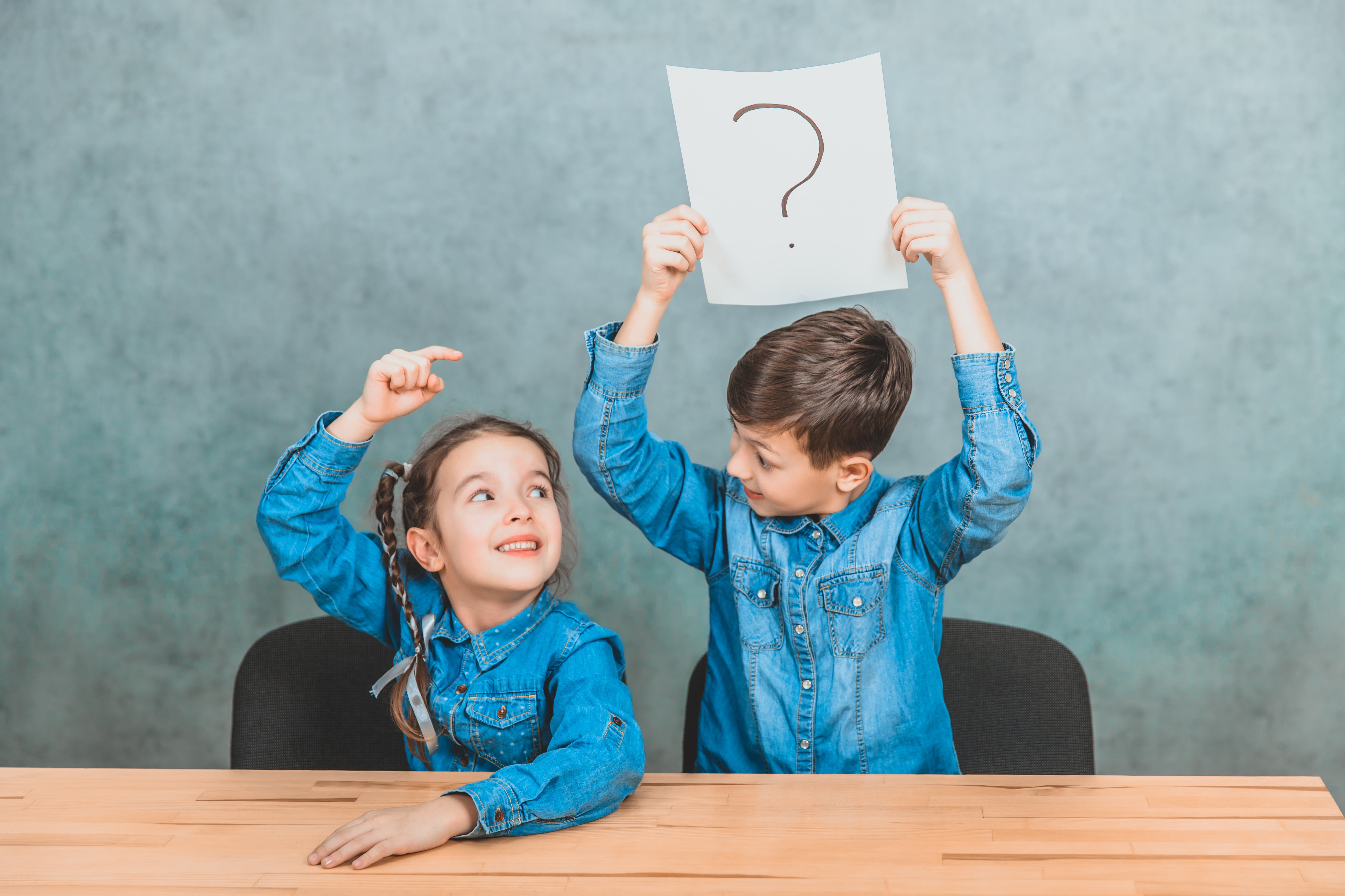 Schulkinder sitzen am Schreibtisch. Ein Junge hebt ein Blatt Papier mit einem Ausrufezeichen hoch, ein Mädchen schaut nach oben, zeigt mit dem Finger darauf und lächelt.